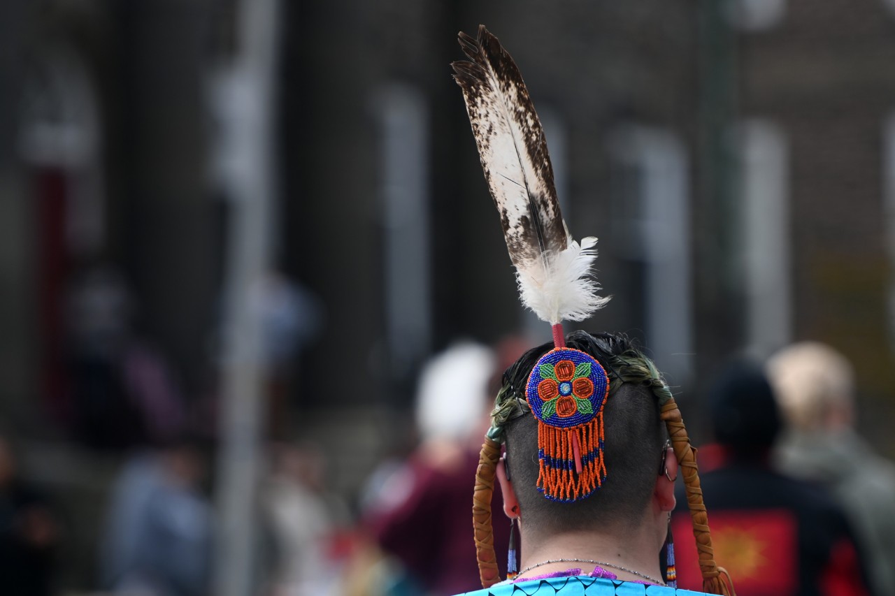 The back of a person wearing a traditional Mi'Kmaq headdress with beading and feather. Mawio'mi guests are blurred in the background.