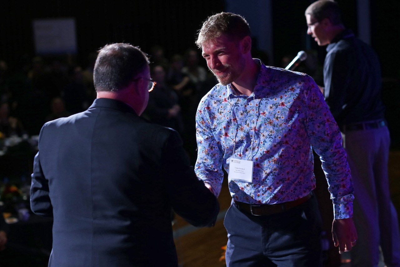 A person wearing a suit, with short grey hair shakes the hands of a person in a floral shirt with short blond hair.
