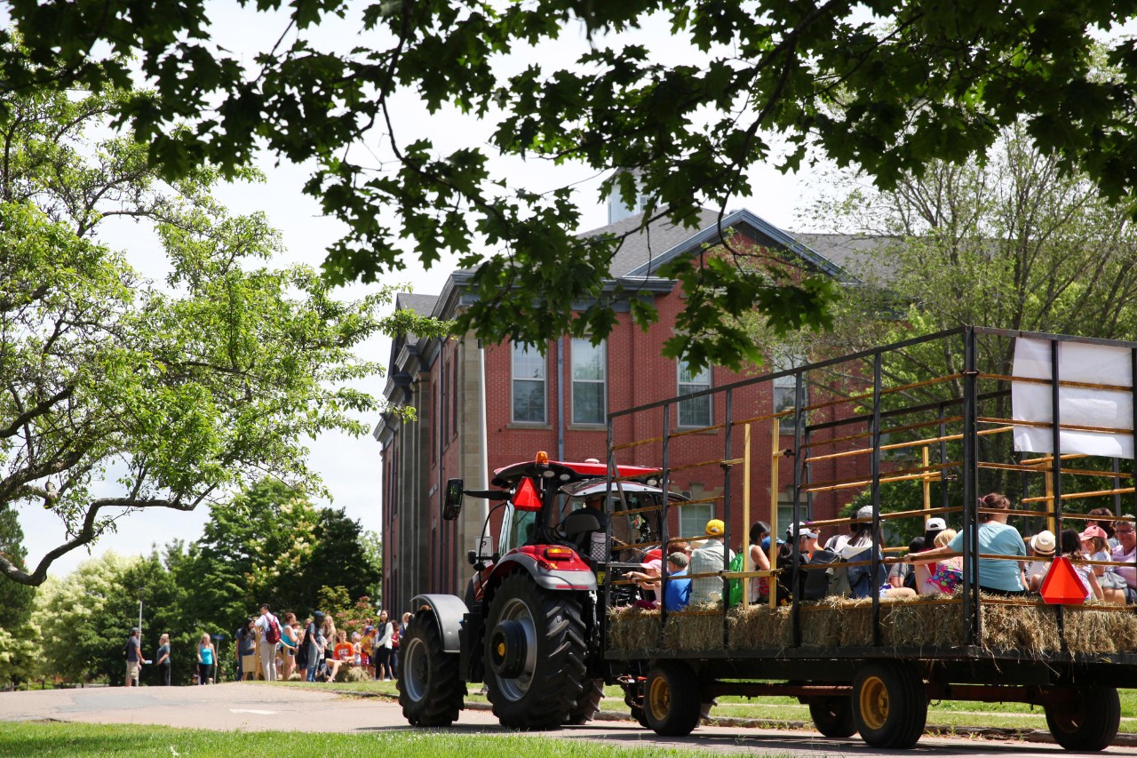 A red tractor pulling a flatbed filled with guests sitting on hay bales approaches a brick building.