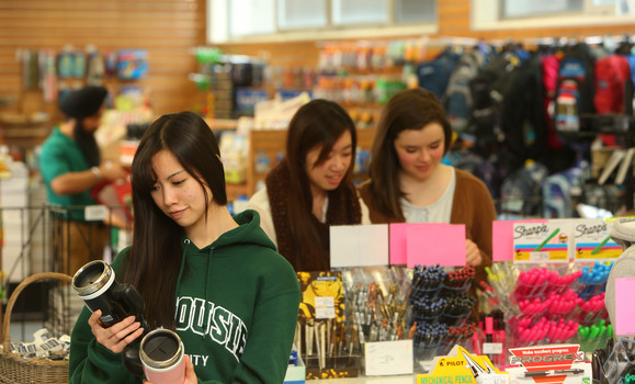 Four students shopping at the Dal Bookstore. One student is in front wearing a green Dalhousie hoodie and holding up two travel mugs. 