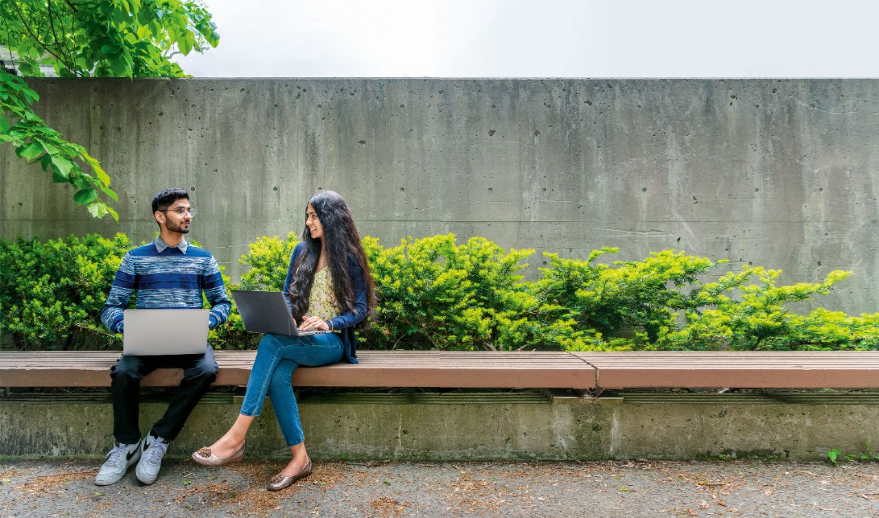 Two students with open laptops sit on a bench in front of a concrete wall with open laptops.