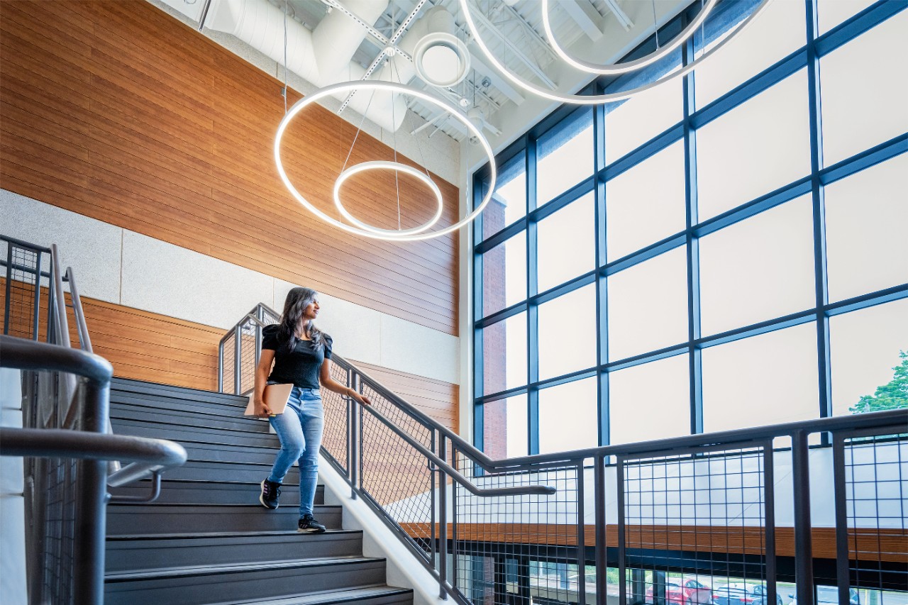 Student holding folder walks down stairs with modern lightfixture and large windows in the background.