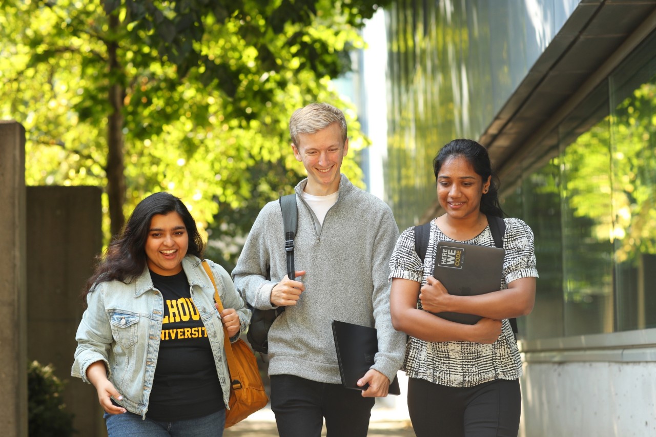 A group of three students walking down a sidewalk with backpacks and laptops.