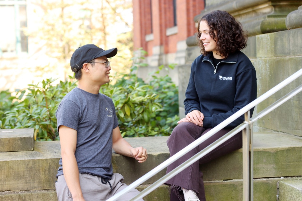 A female-presenting person is sitting on the exterior stone steps of the Dal Architecture building in conversation with a male-presenting person standing and leaning on the side of the steps.