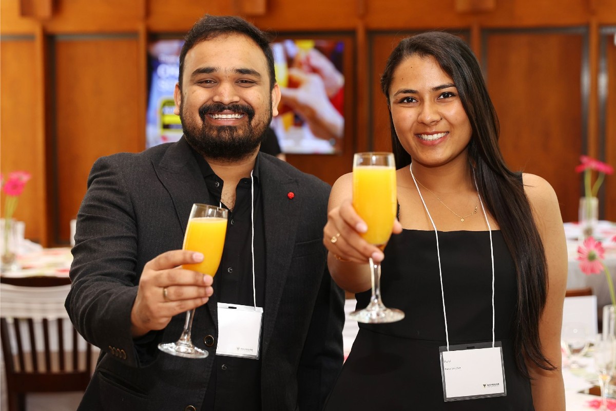 A man and a woman hold out champagne glasses with orange juice gesturing as a toast while at an event in weekend brunch attire.