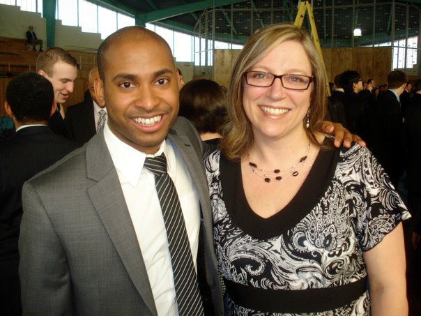 A man wearing a grey suit and white shirt poses with his arm around a woman wearing glasses and a black and white shirt