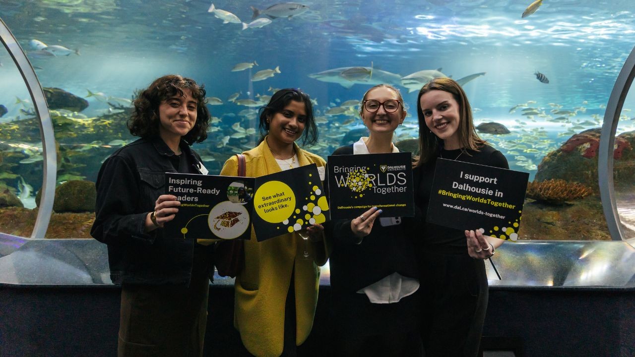 Four recent alumni hold Dal signs standing in front of the aquarium with aquatic species swimming around.