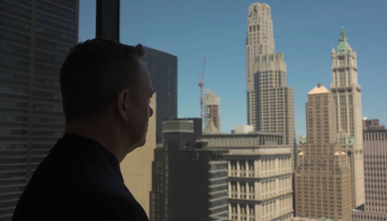 Langlands is looking out a window at a city scape of high-rise buildings.