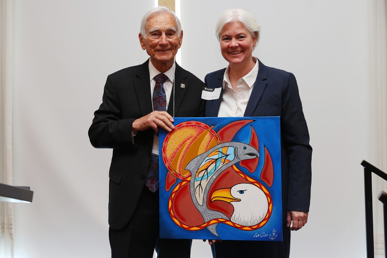 Two people pose holding a painting at an event.