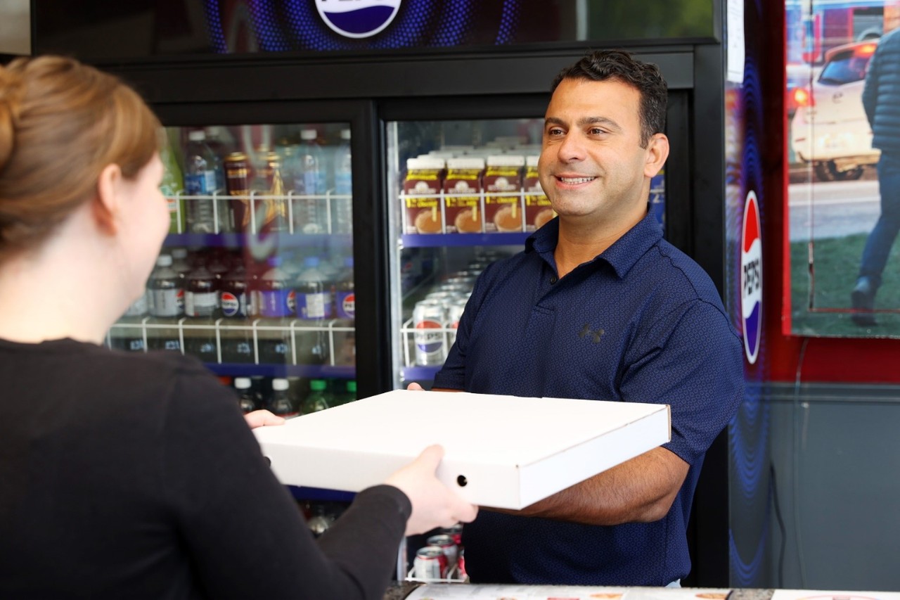 A man is smiling and handing a pizza box to someone across the restaurant counter.