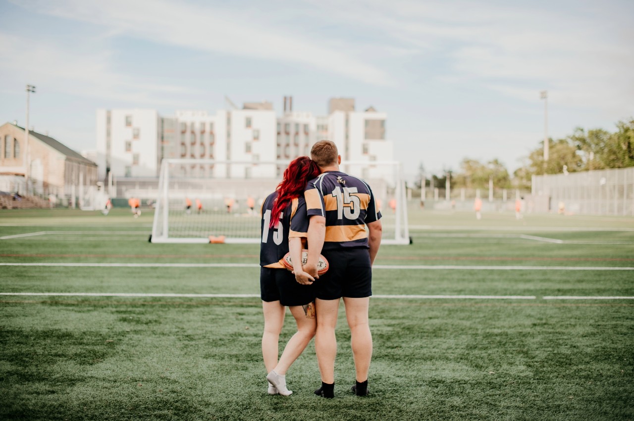 A man and woman hold a rugby ball together behind them overlooking a sports field.