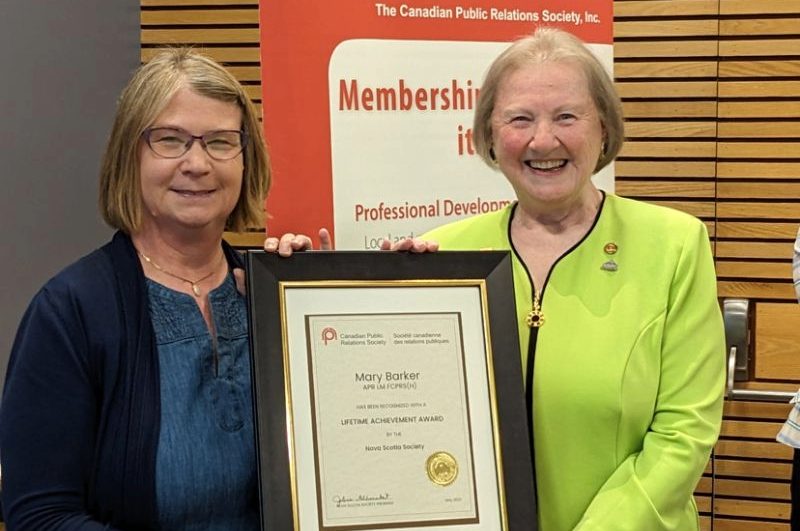 Barker is smiling and holding a framed certificate of her award alongside another women in front of a banner at an event.