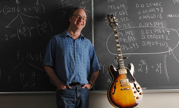 Dr. Jason Brown with his guitar in a classroom with equations on a chalkboard