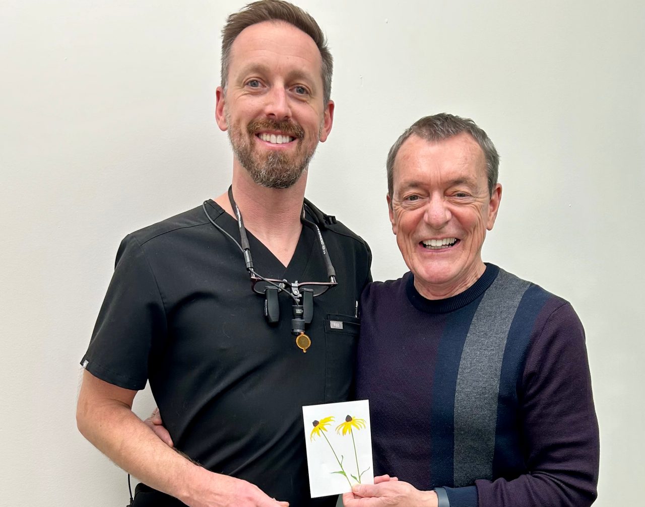 A man wearing black scrubs stands alongside a man in a purple sweater who is holding a card with two yellow yellow flowers.