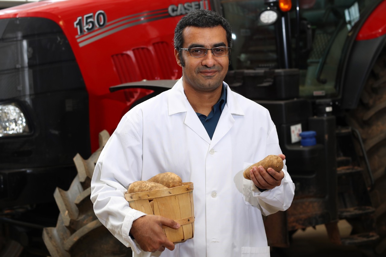A mane wearing glasses and a white lab coat stands in front of a tractor holding a basket of potatoes.