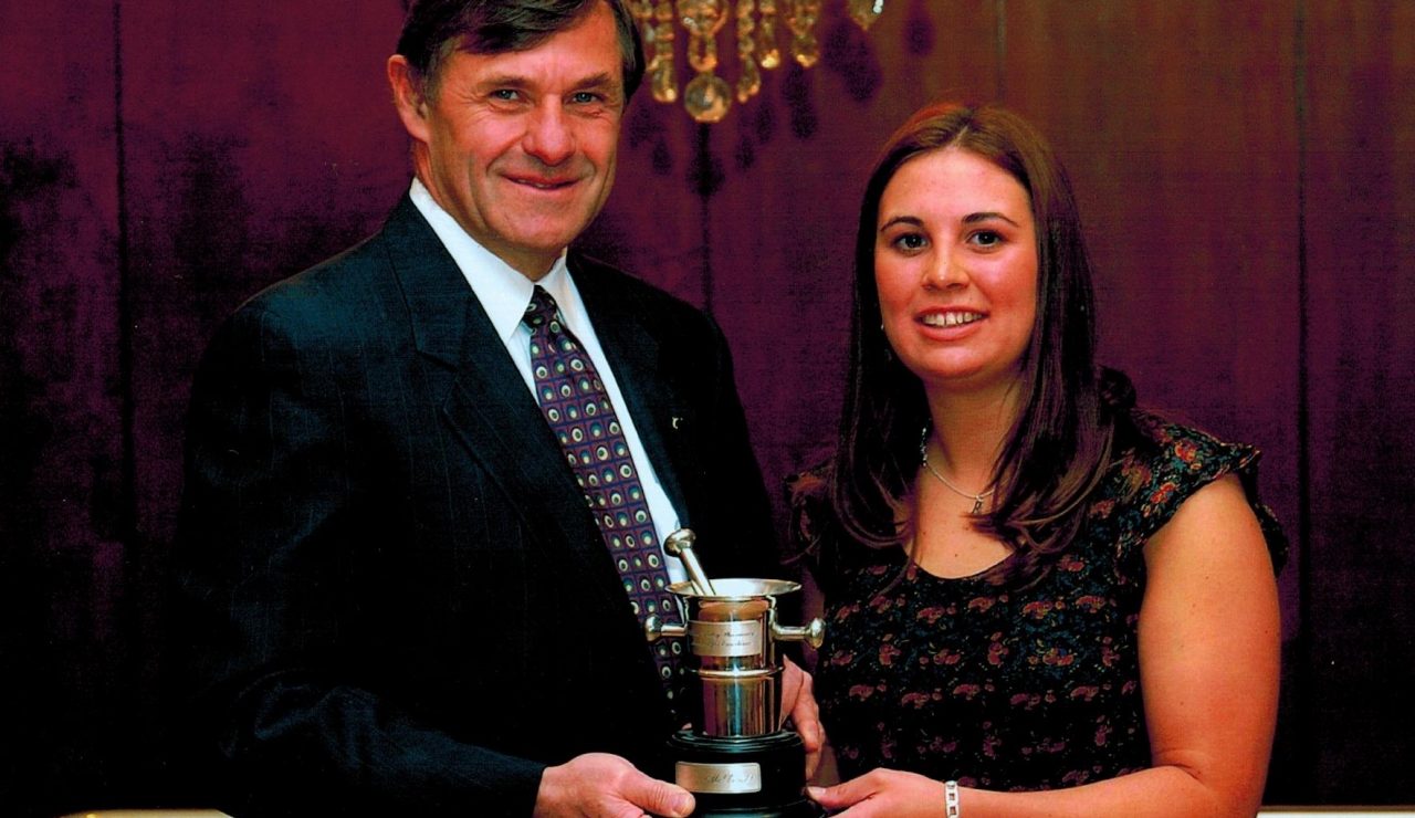A man wearing a suit and tie holds a small trophy with a woman wearing a floral print blouse