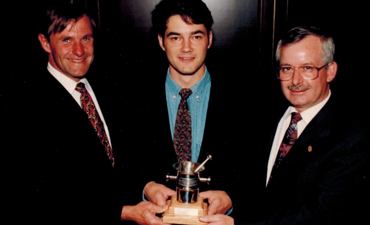 Three men wearing suits hold an award together.