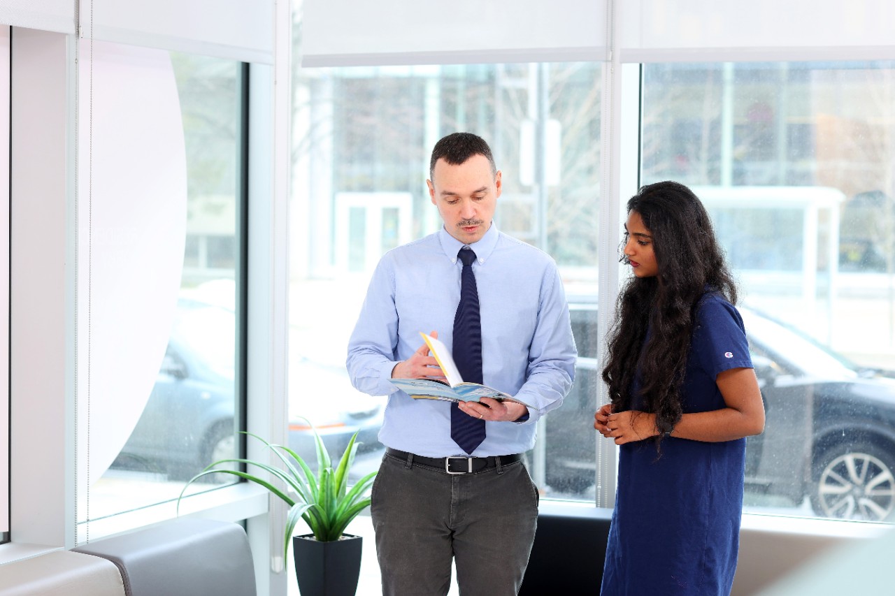 A male-presenting advisor wearing a tie shows a brochure to a female-presenting student who looks on.