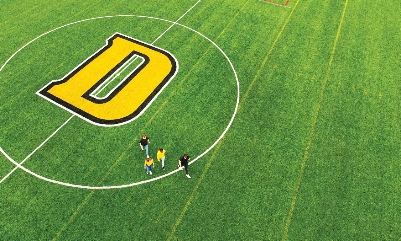 Aerial view of four students wearing black and yellow crossing green sports field painted with a large D surrounded by a white circle.